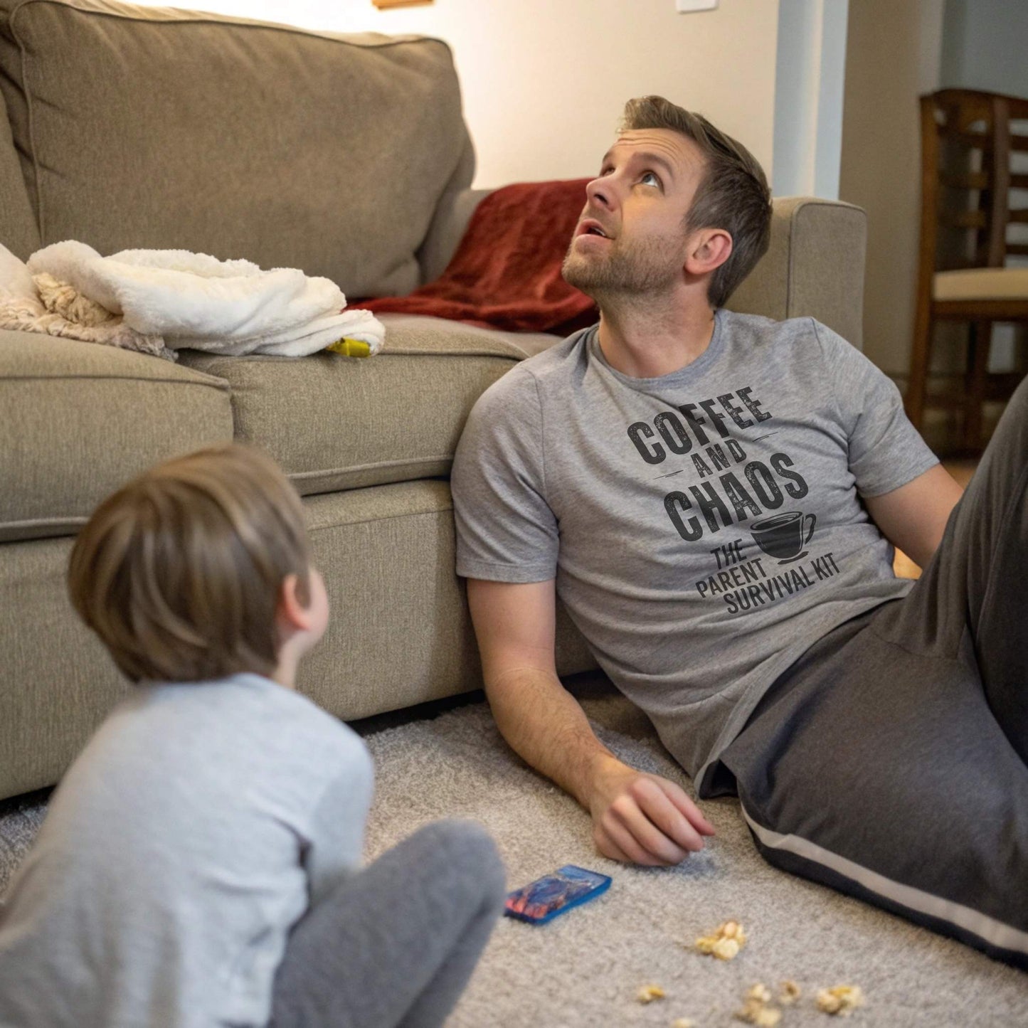 A dad in an Athletic Heather "Coffee and Chaos" t-shirt, looking up with a child on the floor, capturing the humorous chaos of parent life.