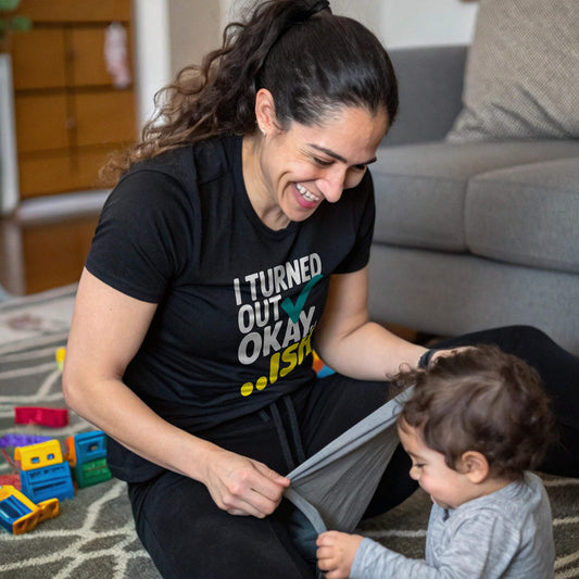 Mom in a black "I Turned Out Okay...ish" t-shirt, laughing joyfully while playing with her baby on the floor, showcasing mom life humor.