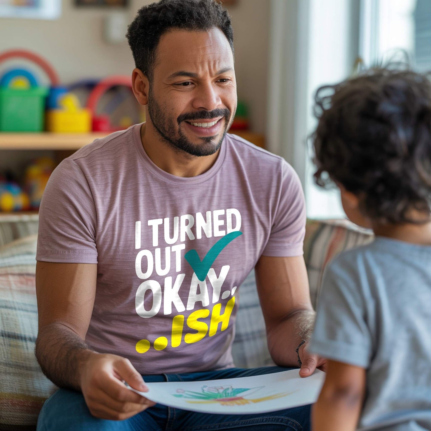 Dad in a Heather Mauve "I Turned Out Okay...ish" t-shirt, smiling at his child while looking at a drawing, depicting a heartwarming parent moment.
