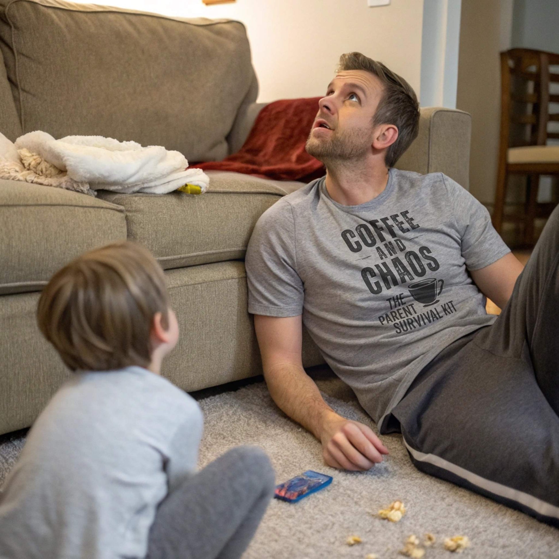 A dad in an Athletic Heather "Coffee and Chaos" t-shirt, looking up with a child on the floor, capturing the humorous chaos of parent life.