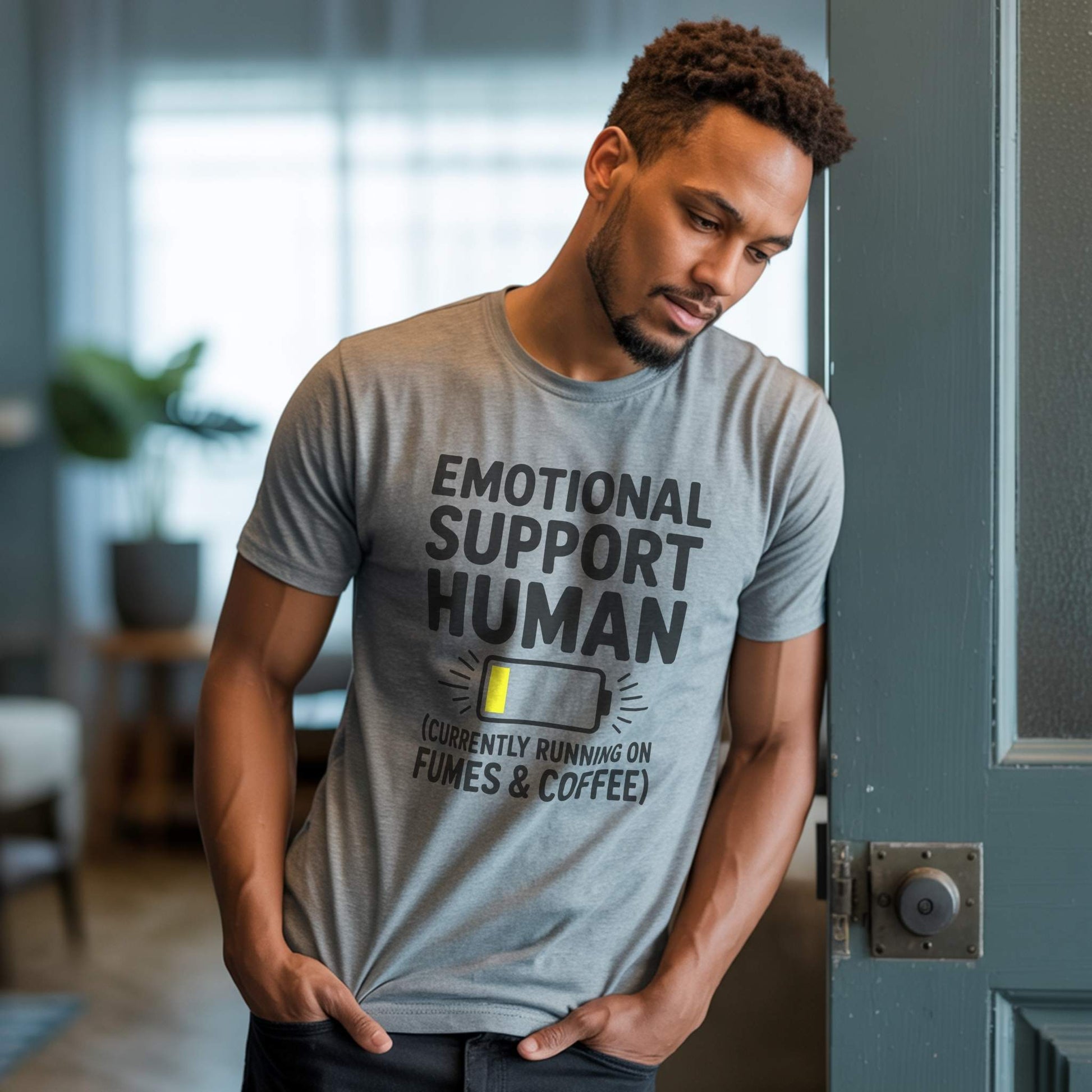 ifestyle Photo - Parent; Male parent wearing an Athletic Heather "Emotional Support Human" t-shirt, leaning against a doorframe, showing a moment of quiet parent reflection.