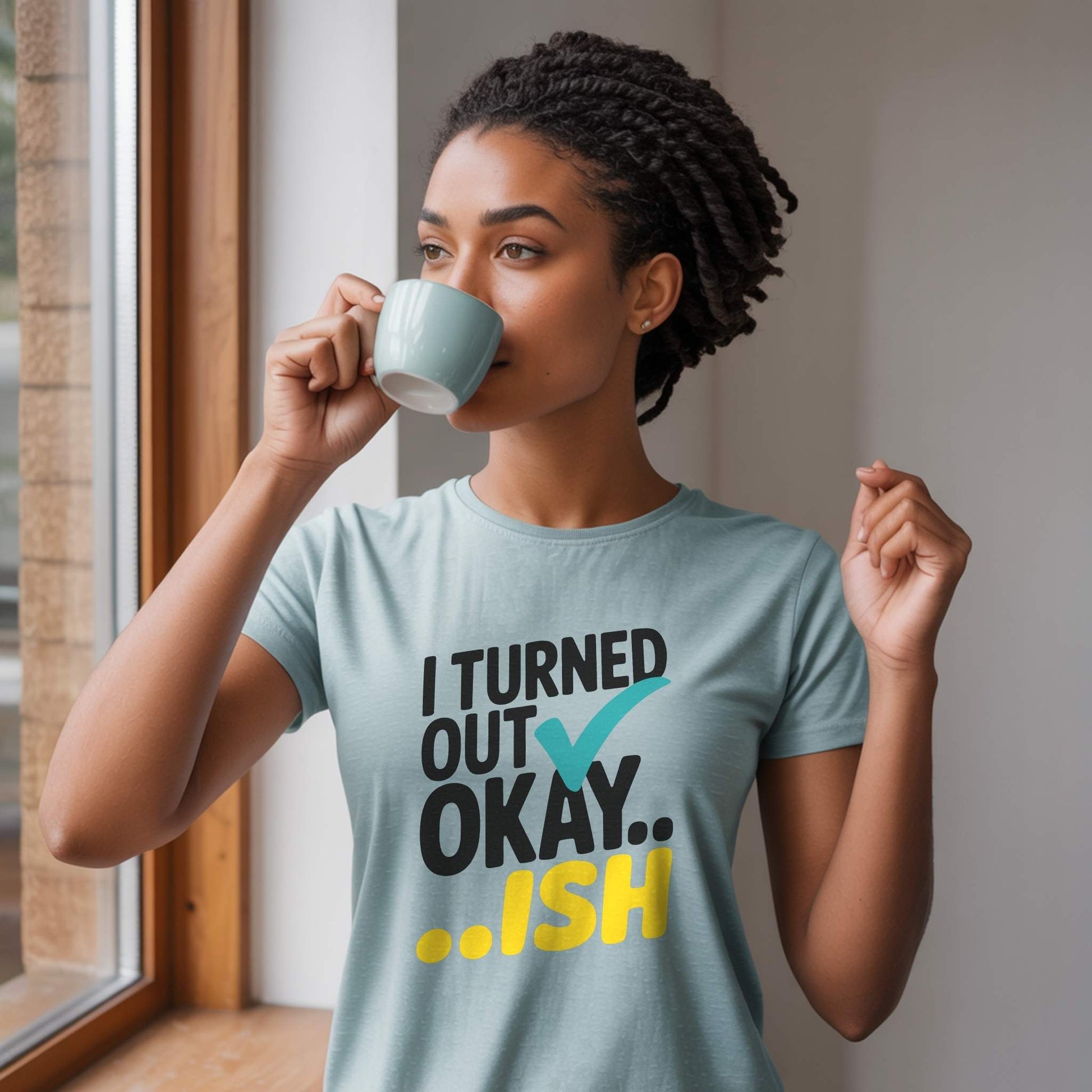 Woman in a Heather Prism Dusty Blue "I Turned Out Okay...ish" t-shirt, sipping coffee by a window, enjoying a moment of self-care.