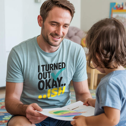 Dad in a Heather Prism Dusty Blue "I Turned Out Okay...ish" t-shirt, happily looking at his child's drawing, embracing parenting realness.