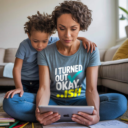 Mom in a Heather Slate "I Turned Out Okay...ish" t-shirt, sitting on the floor with her child looking at a tablet, portraying busy mom life.