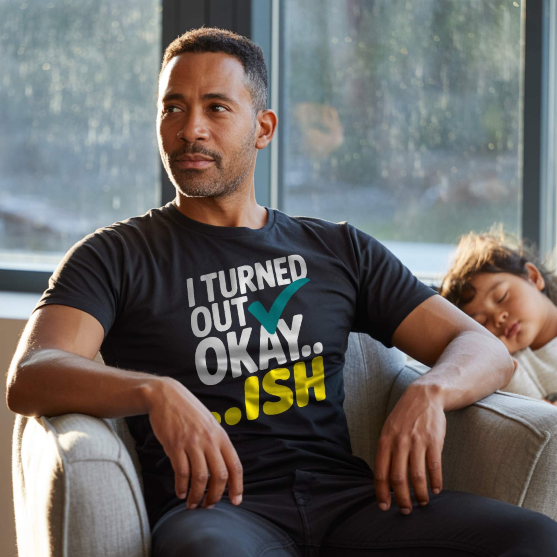 Dad in a black "I Turned Out Okay...ish" t-shirt, looking thoughtful with a child sleeping next to him, representing relatable parent struggles.