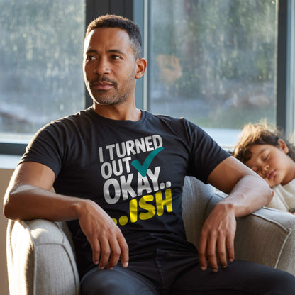 Dad in a black "I Turned Out Okay...ish" t-shirt, looking thoughtful with a child sleeping next to him, representing relatable parent struggles.