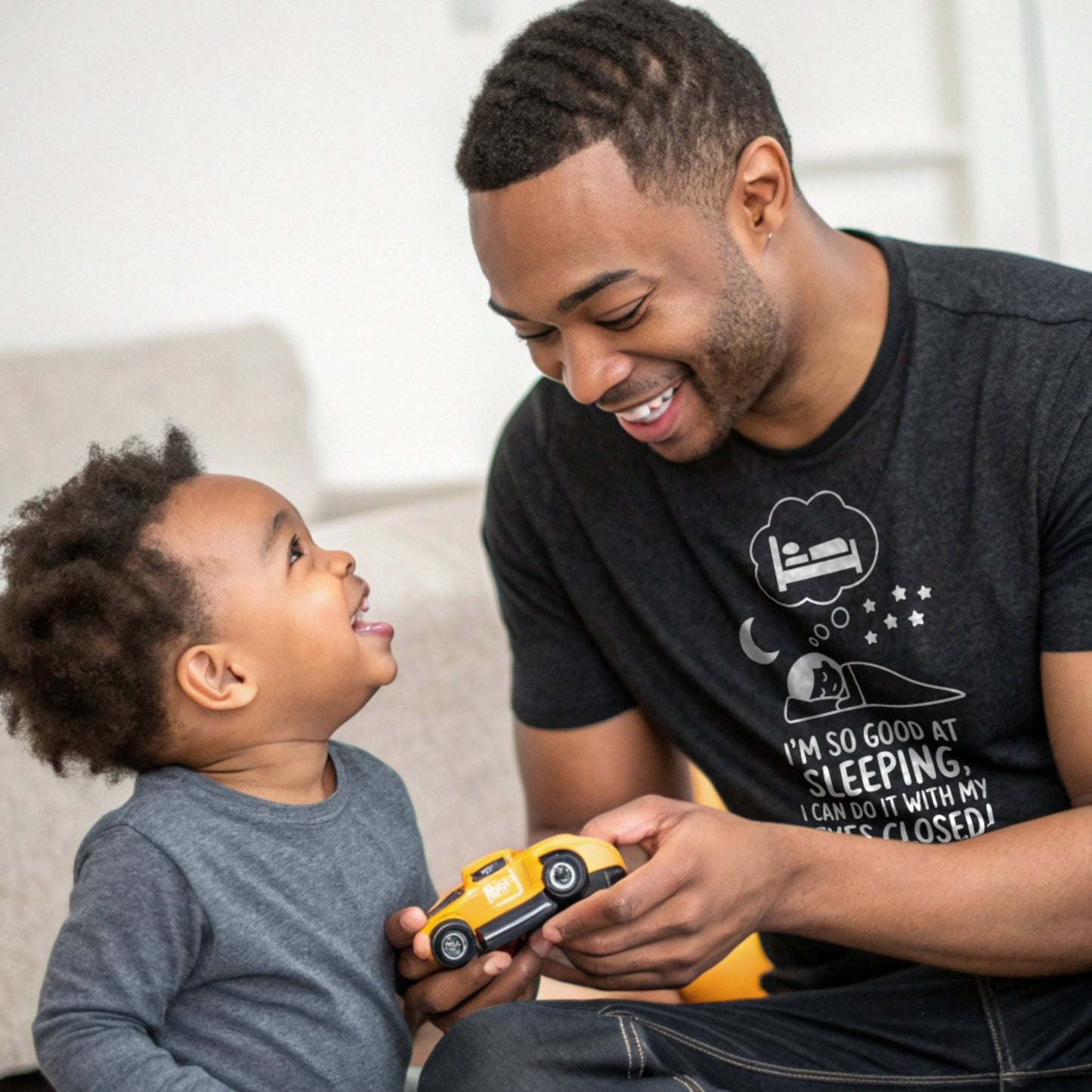 Father and child playing, dad in a heather black "I'm so good at sleeping" t-shirt, showing dad life and shared joy. 