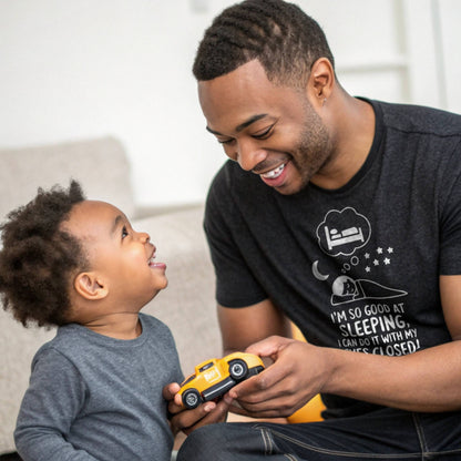 Father and child playing, dad in a heather black "I'm so good at sleeping" t-shirt, showing dad life and shared joy. 