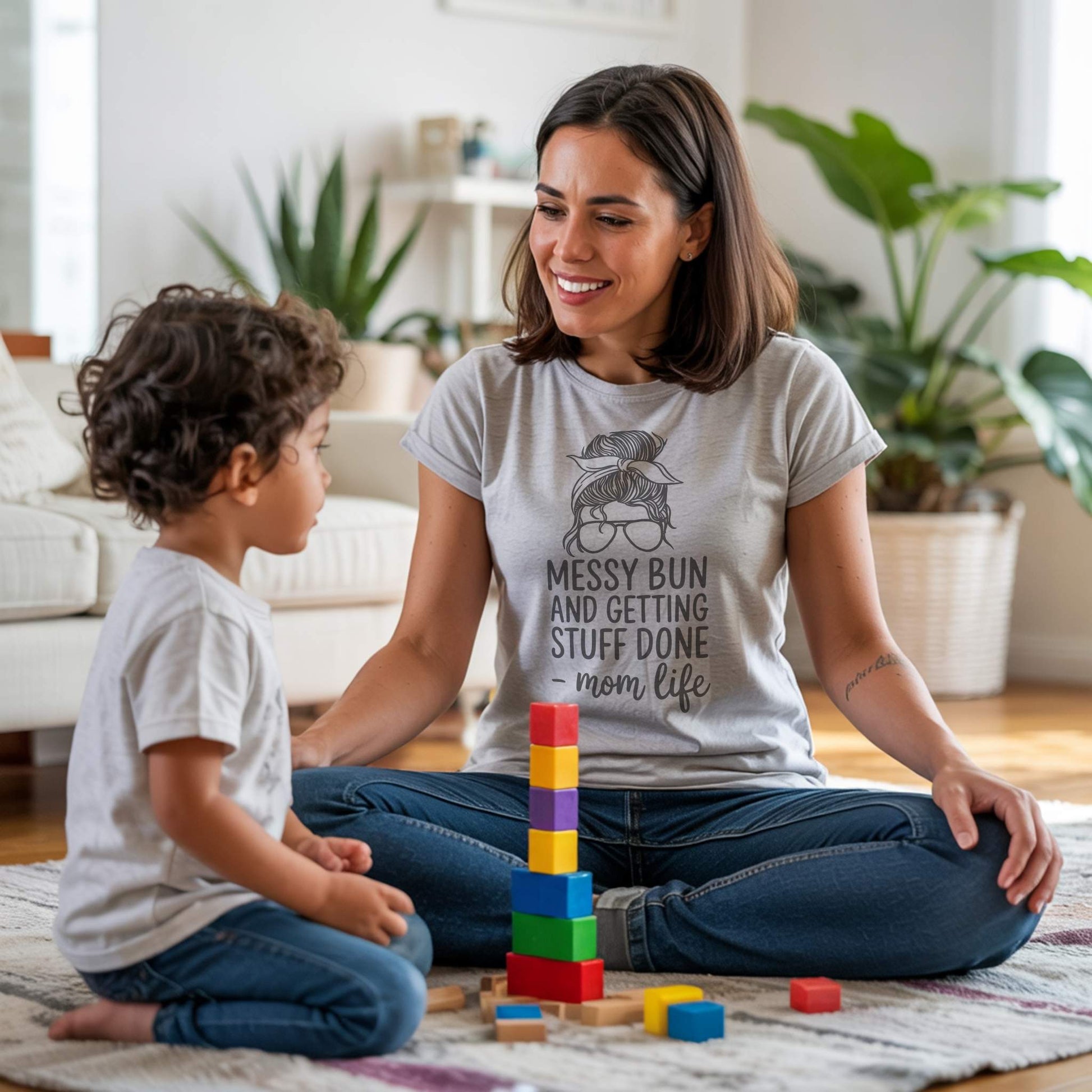 Mom in Athletic Heather "Messy Bun" t-shirt playing blocks with child, showing relatable parent life.