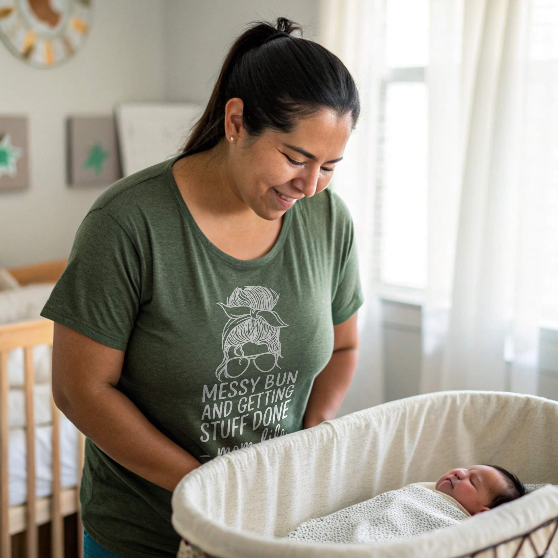 New mom in Heather Forest "Messy Bun" t-shirt gently looking at baby in bassinet, a comforting parent moment.