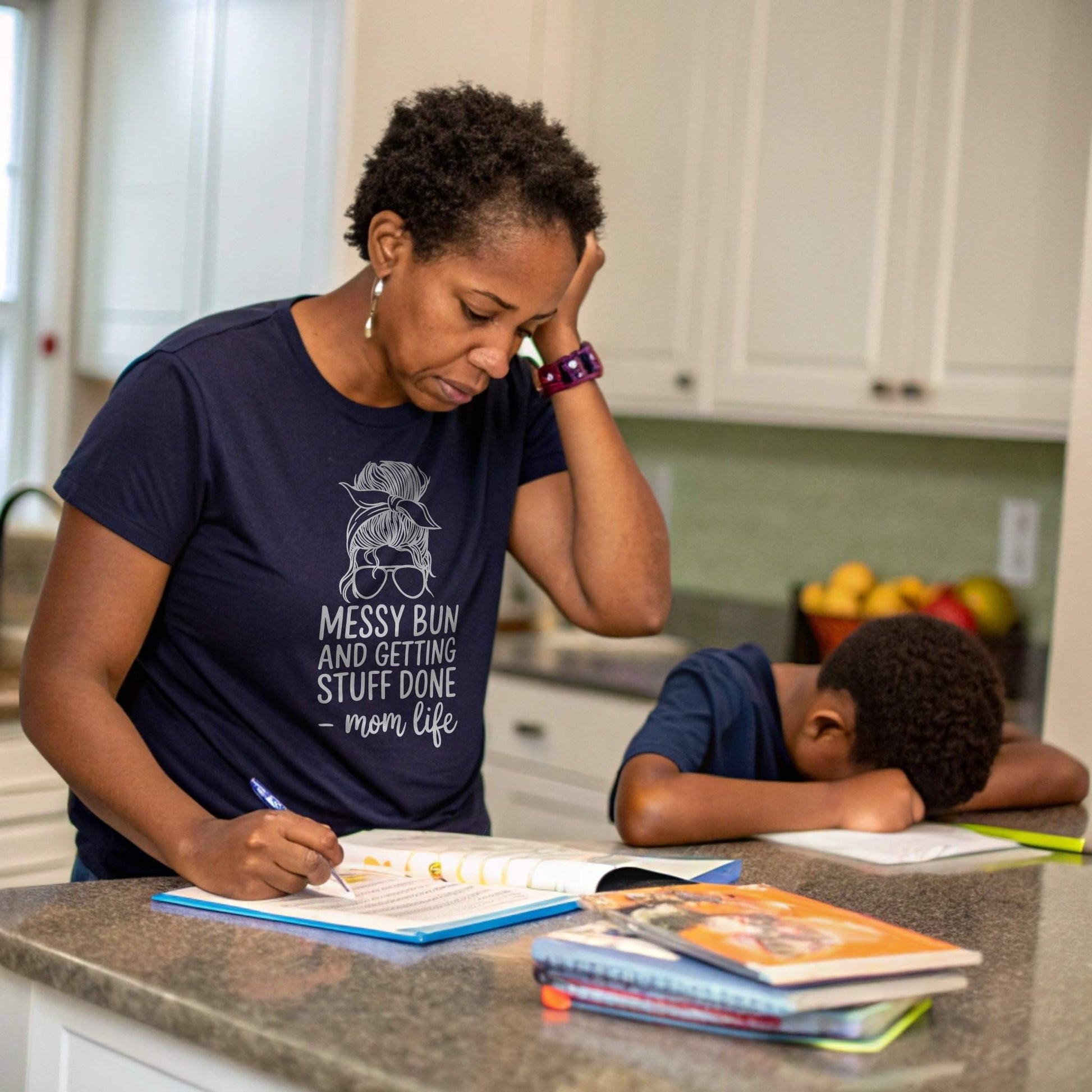 Stressed mom in Navy "Messy Bun" t-shirt doing homework with child, capturing real parent struggles.